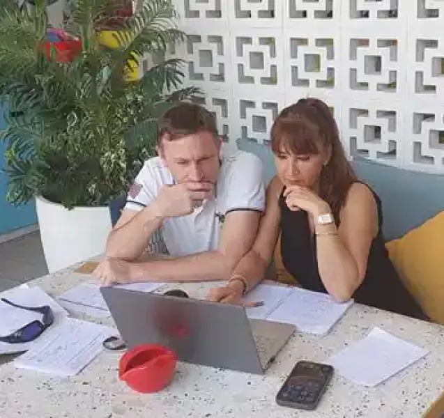 Photo de Jean-Christophe et Nathalie concentrés devant un ordinateur portable, entourés de documents sur une table en extérieur. Ils travaillent ensemble dans un cadre agréable, décoré de plantes et d’un mur à motifs modernes, reflétant une ambiance studieuse et conviviale.